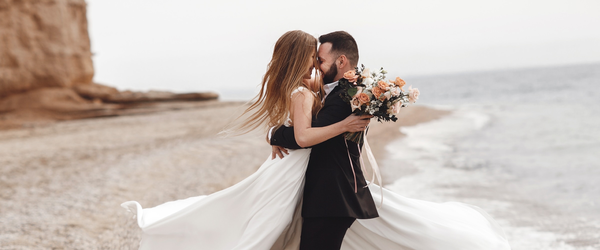 Photo of just married couple embracing at the water's edge of a beach.