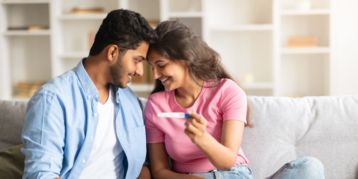 Photo of Indian couple on couch looking at each other and smiling over pregnancy test.