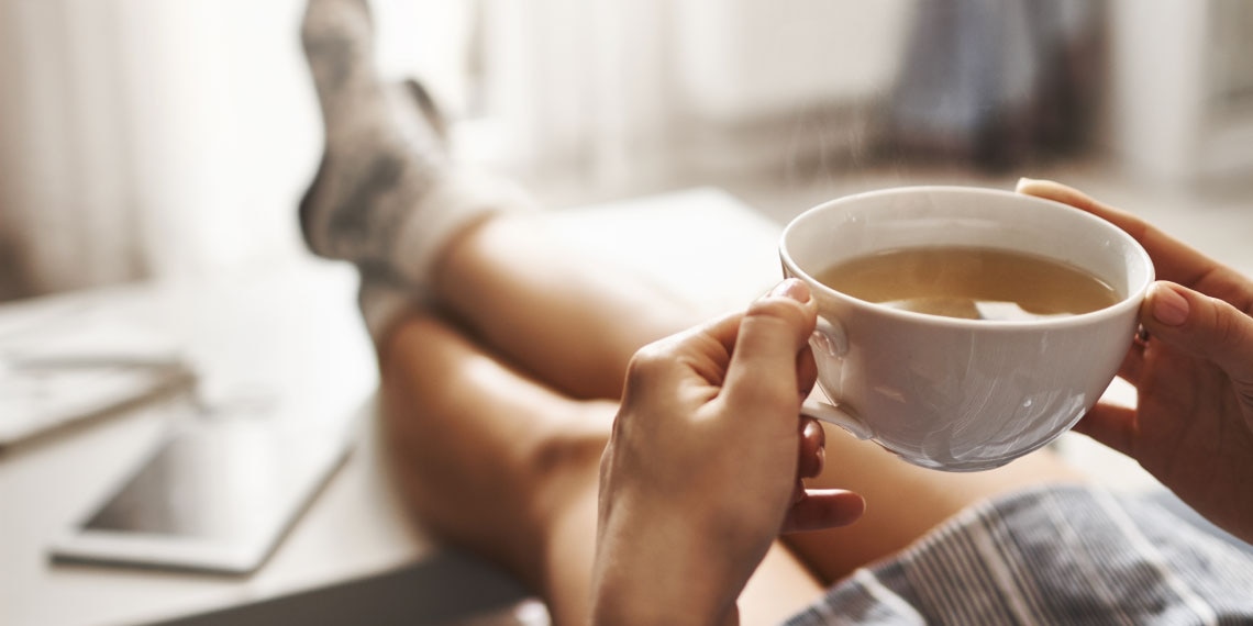 Photo of legs propped up on an ottoman and hands are holding a coffee cup.