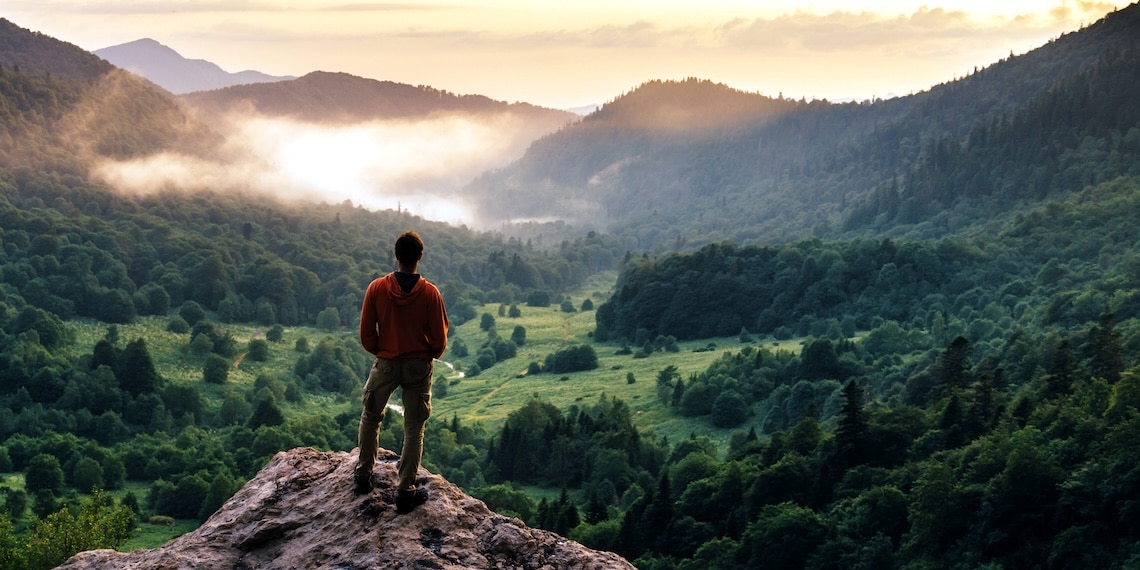 Photo of young man standing on a rock peak with view of valley below him.