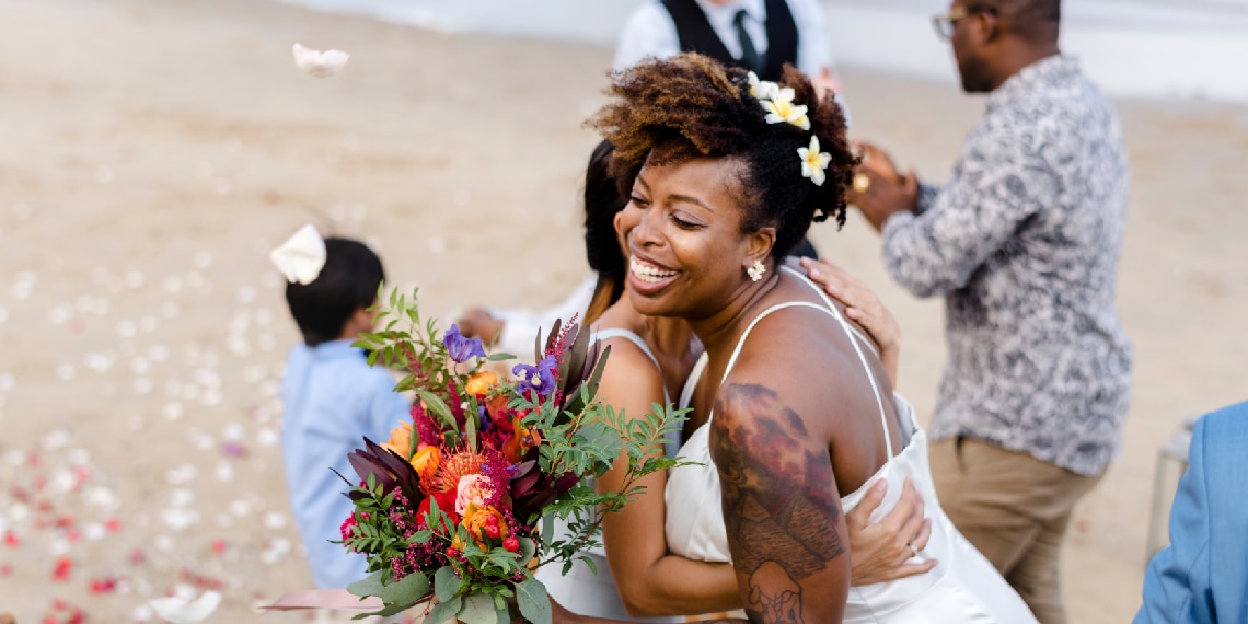 Photo of woman hugging African American bride on the beach.